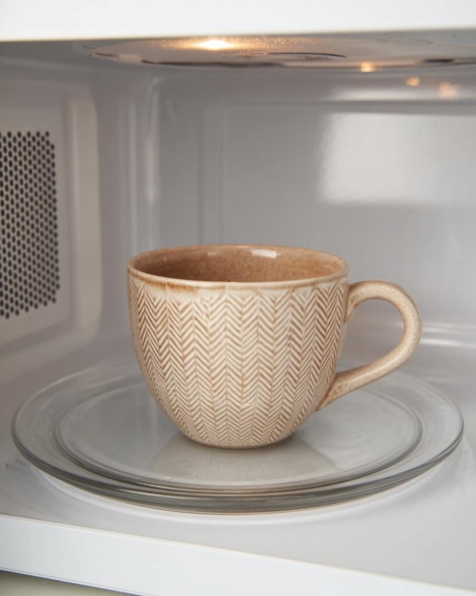 A light brown ceramic cup with a handle sits in the middle of a microwave's glass turntable. The cup has a repeating herringbone pattern etched on the outside, giving it a textured look. The microwave interior is clean and white with small vent holes on the sides and back, and the inside walls have a smooth finish. The glass turntable is clear and circular with a slight raised edge. The scene is simple and bright, showing the cup ready to be warmed. photo taken with an iphone --ar 4:5 --v 7