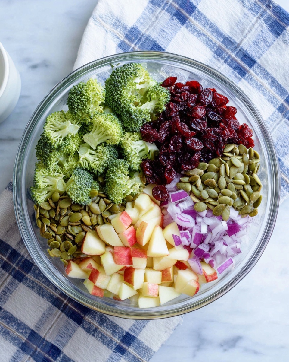 A clear glass bowl shows five sections of fresh ingredients on top of a white marbled surface with a blue and white checkered cloth underneath. The bowl contains bright green broccoli florets, small dark red dried cranberries, light green pumpkin seeds, reddish-white chopped apple pieces, and finely chopped purple onions. Each ingredient stays separate in its own area, making a colorful and fresh look. A white cup is slightly visible on the side. Photo taken with an iphone --ar 4:5 --v 7