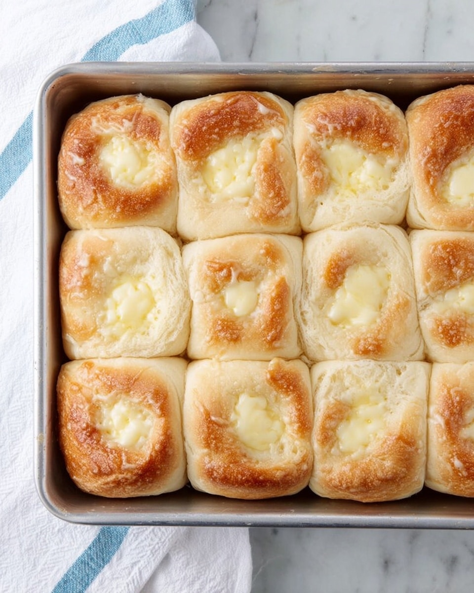 The image shows nine soft, golden-brown rolls arranged in a 3x3 grid inside a silver baking pan. Each roll has a visible creamy, pale yellow layer of cheese or custard on top, nestled in the center and slightly melted. The rolls have a light, fluffy texture with a shiny, slightly crusty surface. The pan is placed on a white marbled surface with a white towel with blue stripes partially visible on the left side. photo taken with an iphone --ar 4:5 --v 7