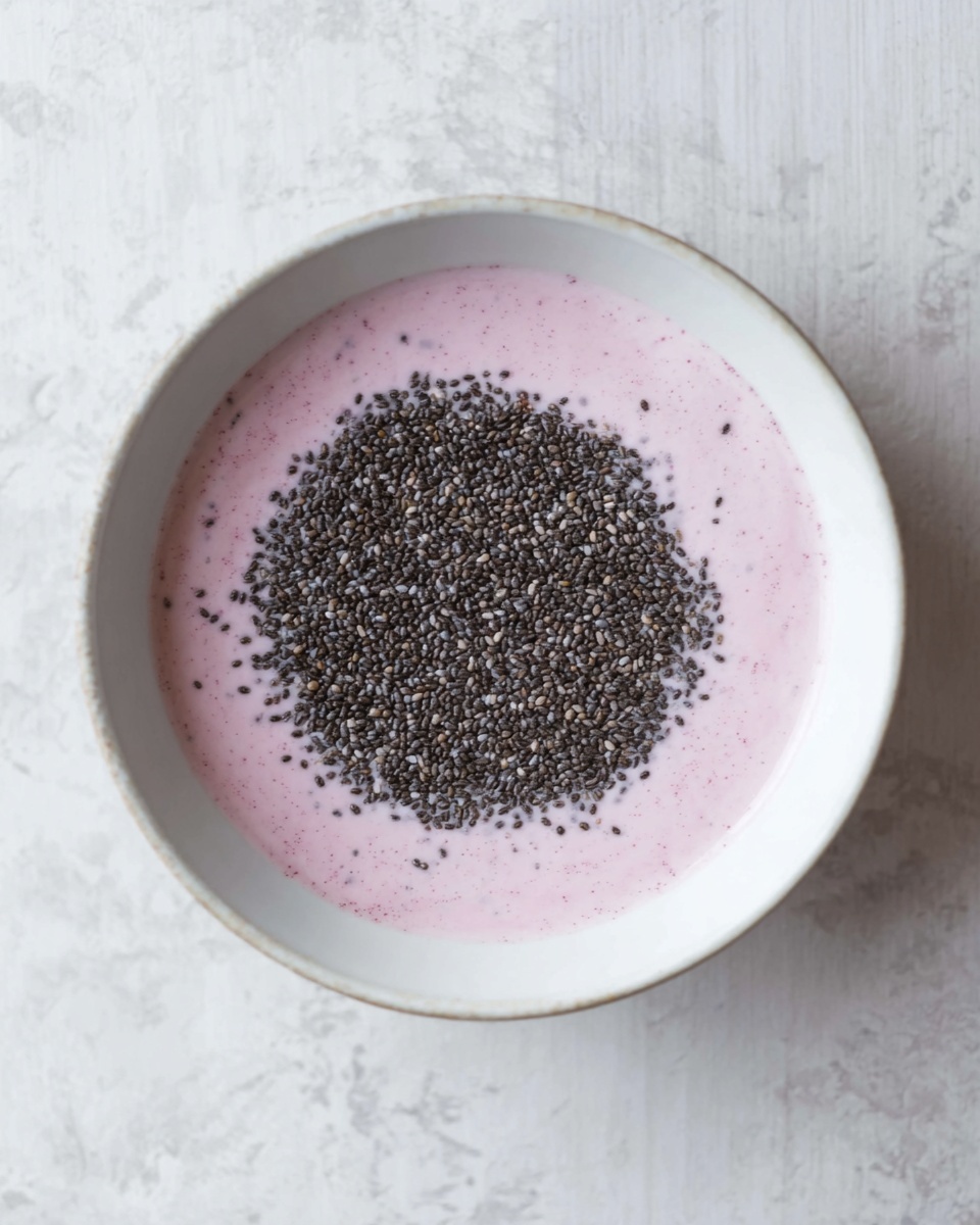 A white bowl with a smooth light pink yogurt-like base fills the entire bowl. In the center of this base is a round pile of small black chia seeds, densely packed and slightly raised above the yogurt surface. The bowl sits on a white marbled textured surface. The lighting is soft and natural, showing clear textures in the seeds and yogurt. Photo taken with an iphone --ar 4:5 --v 7