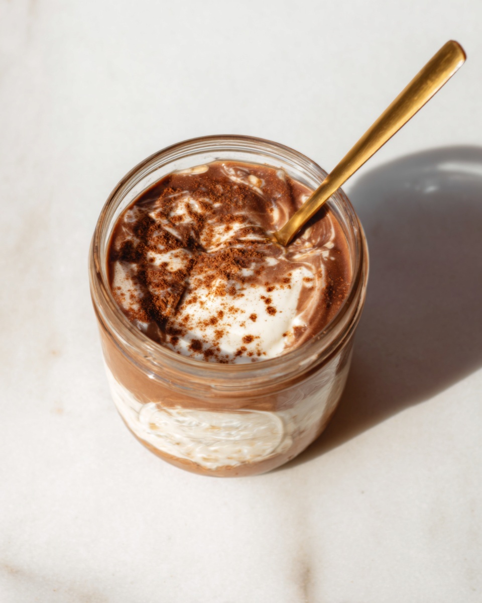 A clear glass jar shows thick layers of creamy white yogurt and rich brown chocolate, swirled together with a light sprinkle of powdered cinnamon or cocoa on top. A gold spoon rests inside the jar, angled towards the upper left, with a woman's hand holding it. The jar sits on a white marbled surface, and soft shadows stretch out from the jar and spoon under bright light. photo taken with an iphone --ar 4:5 --v 7