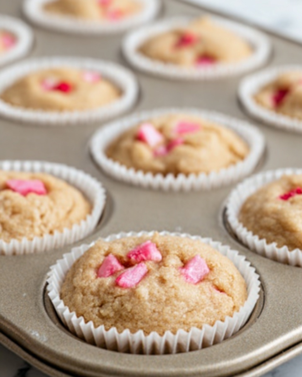 The image shows a close-up of a metal muffin tray holding six soft, light brown cookies with pink chunks embedded on top. Each cookie has a rough, slightly cracked texture and sits inside a white paper cupcake liner, fitting snugly in the tray’s round wells. The photo is focused on the cookie in the bottom left corner, making it the sharpest, while the other cookies blur gradually into the background. The background is a white marbled surface. Photo taken with an iphone --ar 4:5 --v 7