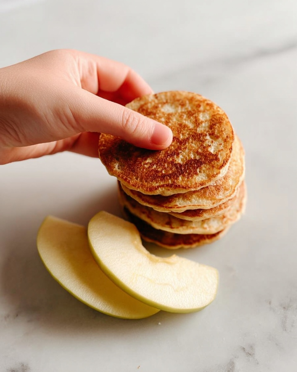 A stack of five small golden-brown pancakes is placed on a white marbled surface, showing a slightly crispy texture on top. Next to the stack are three pale yellow apple slices arranged neatly. A woman's hand is picking the top pancake, pinching it gently between thumb and fingers. The light is soft, highlighting the warm tones of the pancakes and the smooth surface of the apple slices. photo taken with an iphone --ar 4:5 --v 7