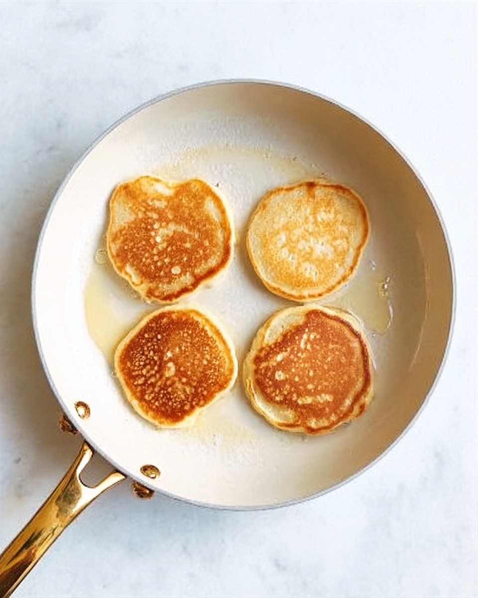 Four small golden-brown pancakes with soft, fluffy texture are in a white frying pan with a gold handle, arranged loosely in a circle. The pancakes have slightly uneven edges with light bubbles visible on top. The frying pan rests on a white marbled surface. Photo taken with an iphone --ar 4:5 --v 7