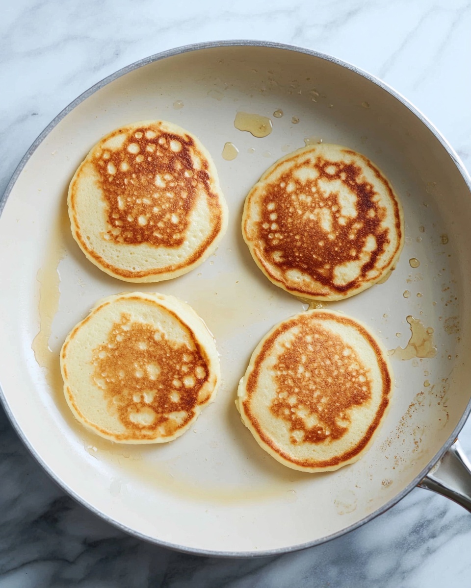 A round white pan holds four small pancakes cooking evenly spaced inside. Each pancake has a light golden brown color with soft, smooth edges and a texture showing tiny bubbles and slight browning patterns on top. The pan surface shows small drops of oil and light marks from cooking. The background is a white marbled surface. photo taken with an iphone --ar 4:5 --v 7