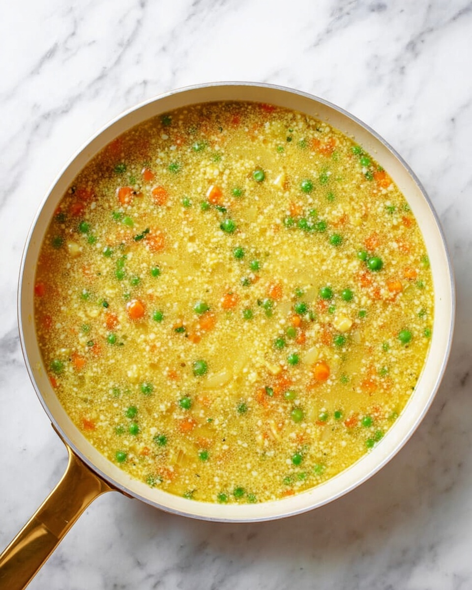 A white pan filled with a mixture of small round yellow grains soaked in broth, with visible small bright green peas and tiny orange carrot pieces scattered evenly throughout. The texture looks soft and wet, with a warm, light yellow color dominating the dish. The pan has a golden handle resting on a white marbled surface, with the food filling the pan almost to the top. photo taken with an iphone --ar 4:5 --v 7