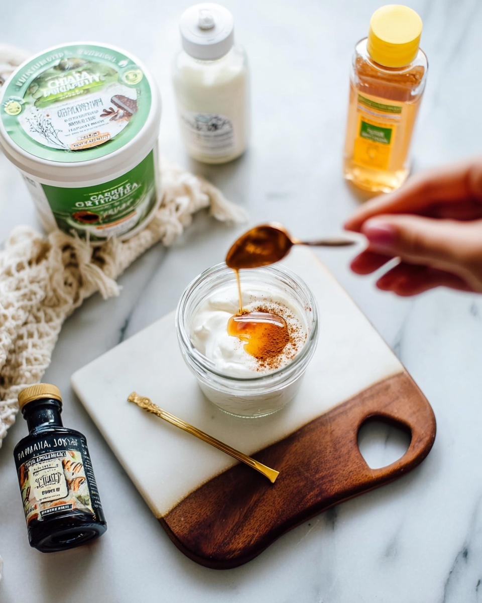 A clear glass jar sits open on a white marble board with a dark wooden edge, filled with white yogurt topped with a small layer of golden honey and a sprinkle of light brown cinnamon being added by a woman's hand holding a cinnamon spice jar. Near the jar is a small gold spoon resting inside. Surrounding the board on the white marbled surface are a white container of plain Greek yogurt with green and black label, a bottle of vanilla extract with a white cap, and a clear honey bottle with a yellow cap. The scene is bright and clean with soft natural light. photo taken with an iphone --ar 4:5 --v 7