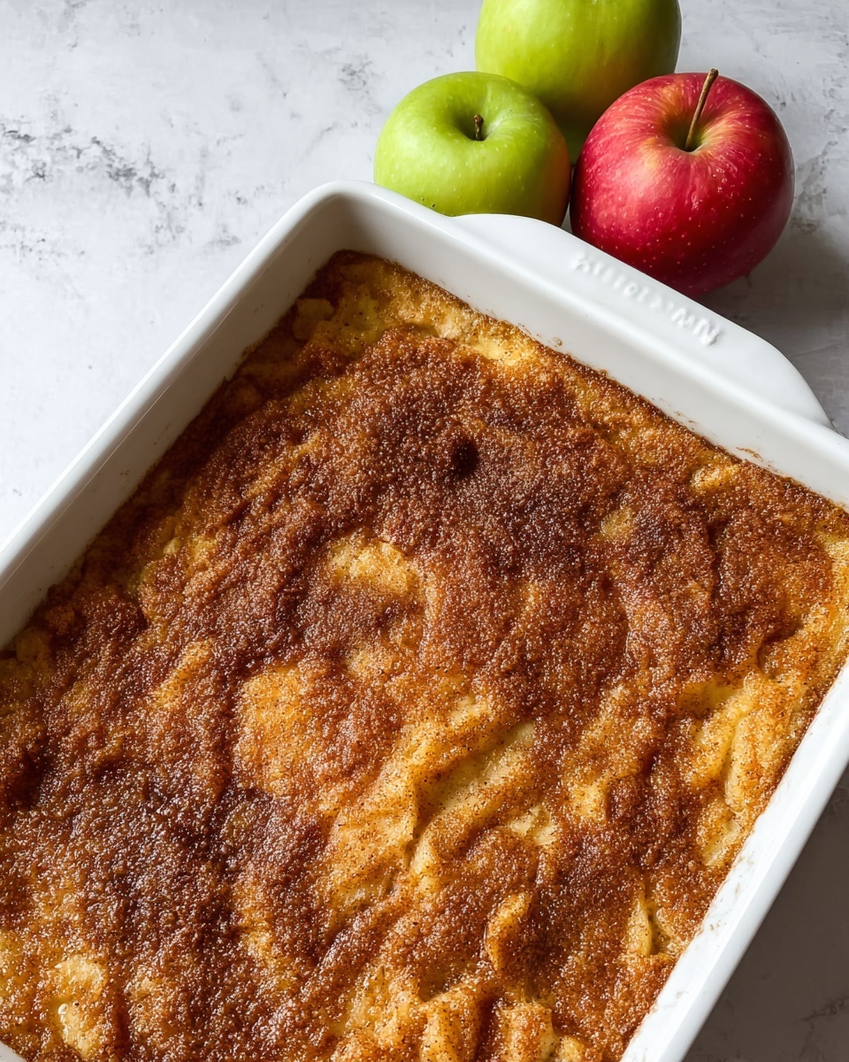 A close-up view of a baked apple dish in a white square ceramic baking dish. The top layer is golden brown with a slightly rough texture, sprinkled evenly with a cinnamon and sugar mixture that adds a darker brown speckled look. The surface shows a mix of soft baked areas and crunchy crust spots, with subtle cracks and uneven ridges. The dish is placed on a white marbled surface, next to three fresh apples showing red and green colors. photo taken with an iphone --ar 4:5 --v 7