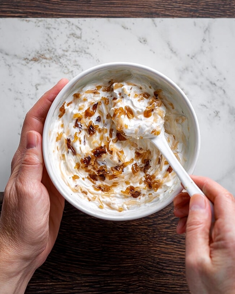 A white bowl held by one woman's hand on the left side and a white spoon held by another woman's hand on the right side, filled with creamy white yogurt mixed with small pieces of golden brown caramelized onions, the mixture showing a textured swirl pattern with bits of onions spread throughout on a dark wooden surface replaced by a white marbled texture background, photo taken with an iphone --ar 4:5 --v 7