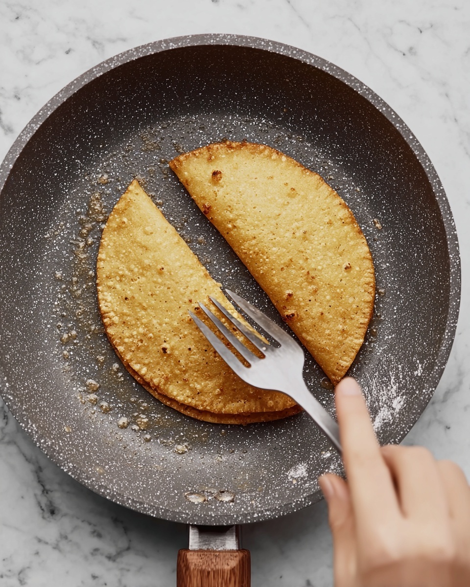 In a dark gray speckled pan with a wooden handle, two golden brown taco shells are shown being fried; one shell is near the top edge of the pan, and the other is being pressed gently with a silver spatula held by a woman's hand in the center of the pan. The taco shells have a crispy texture with small bubbles on the surface. The background is a white marbled texture photo taken with an iphone --ar 4:5 --v 7