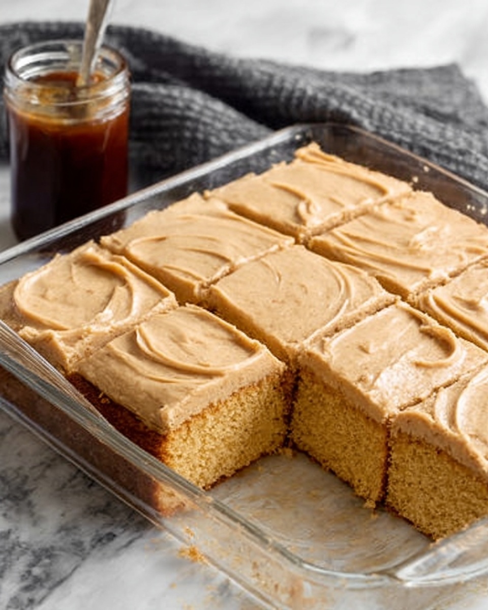 The image shows a glass baking dish with a single layer of thick, soft cake that is golden brown. The cake is cut into six large square slices, with one slice removed from the front. The top of the cake is spread evenly with a smooth layer of light brown frosting that has gentle swirls. Next to the dish is a small glass jar filled with a dark brown sauce. The dish rests on a white marbled surface with a grey cloth in the background. Photo taken with an iphone --ar 4:5 --v 7