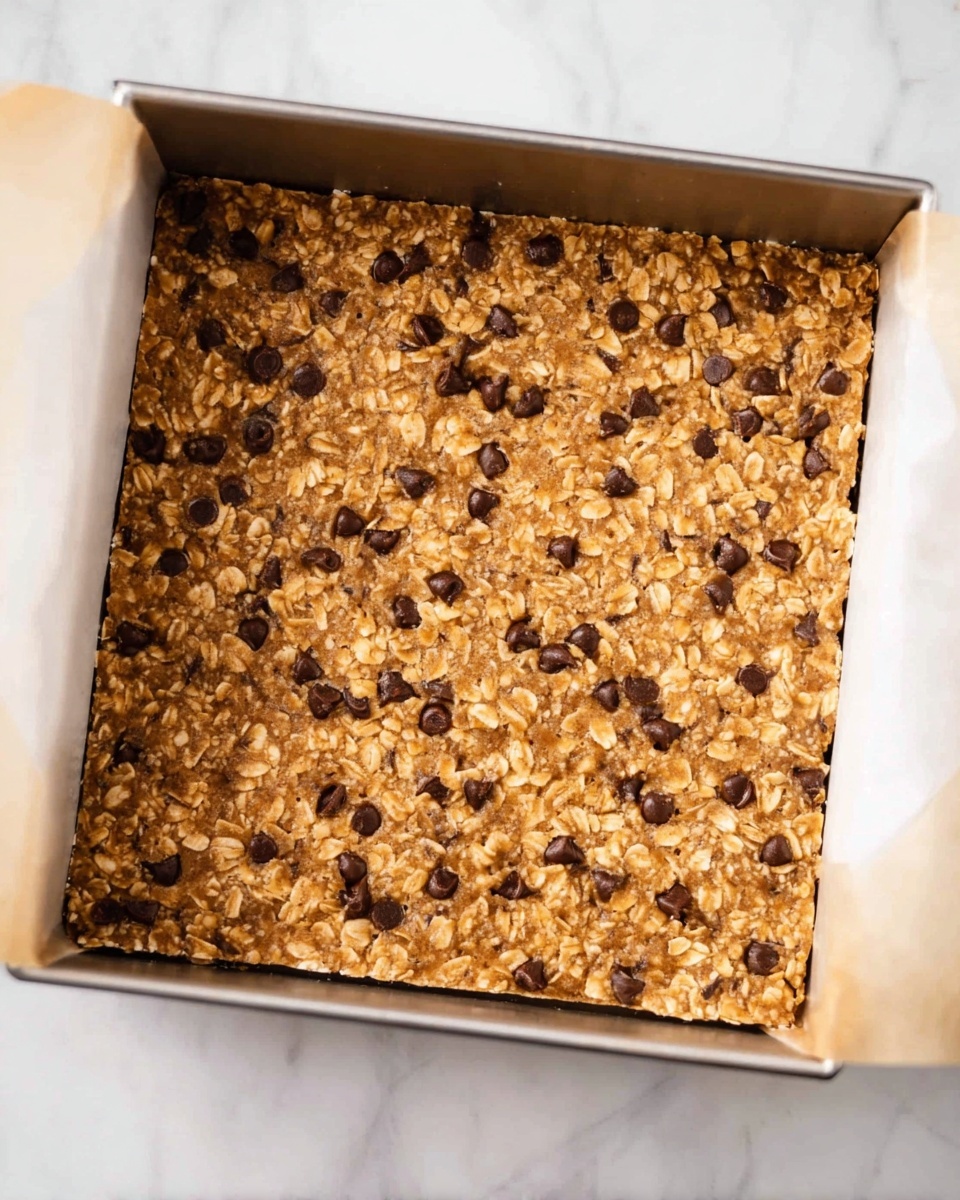 A square baking pan filled with an even layer of golden brown oat and chocolate chip bars, with visible oats and small dark chocolate chips scattered throughout the surface. The pan is lined with parchment paper around the edges, and it is placed on a white marbled surface. The texture of the bars looks slightly soft but firm, showing a homemade, rustic finish photo taken with an iphone --ar 4:5 --v 7
