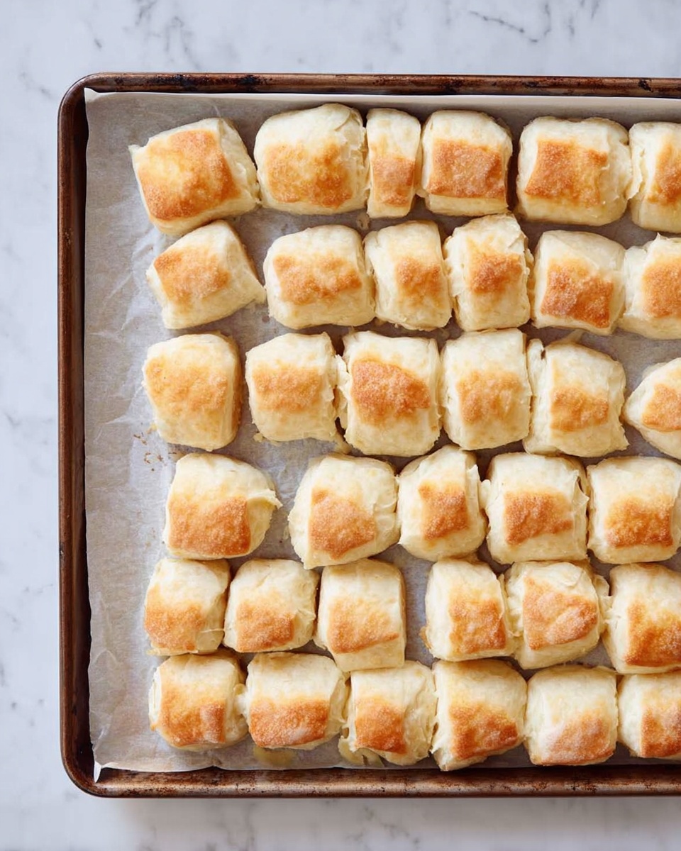 A baking tray filled with small, evenly spaced pieces of baked dough that are golden brown on top and paler on the sides, each piece having a soft, slightly puffed texture with rounded edges. The tray is lined with white parchment paper, and the background surface shows a white marbled texture. The pieces appear light and flaky, arranged in neat rows covering the whole tray. photo taken with an iphone --ar 4:5 --v 7