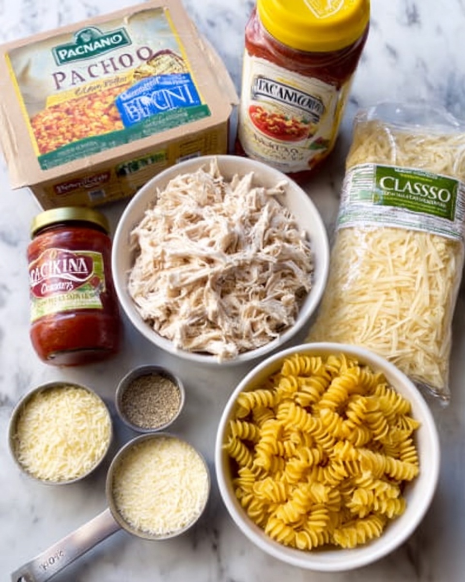 The image shows several cooking ingredients arranged on a white marbled surface. In the front and center, there is a white bowl filled with shredded chicken, to its right another white bowl holds uncooked rotini pasta with a yellow hue. In front of these, a small metal measuring cup is filled with grated parmesan cheese. Around the bowls, various packages and jars are visible: a large box of Panko breadcrumbs on the left, a bottle of spaghetti sauce labeled