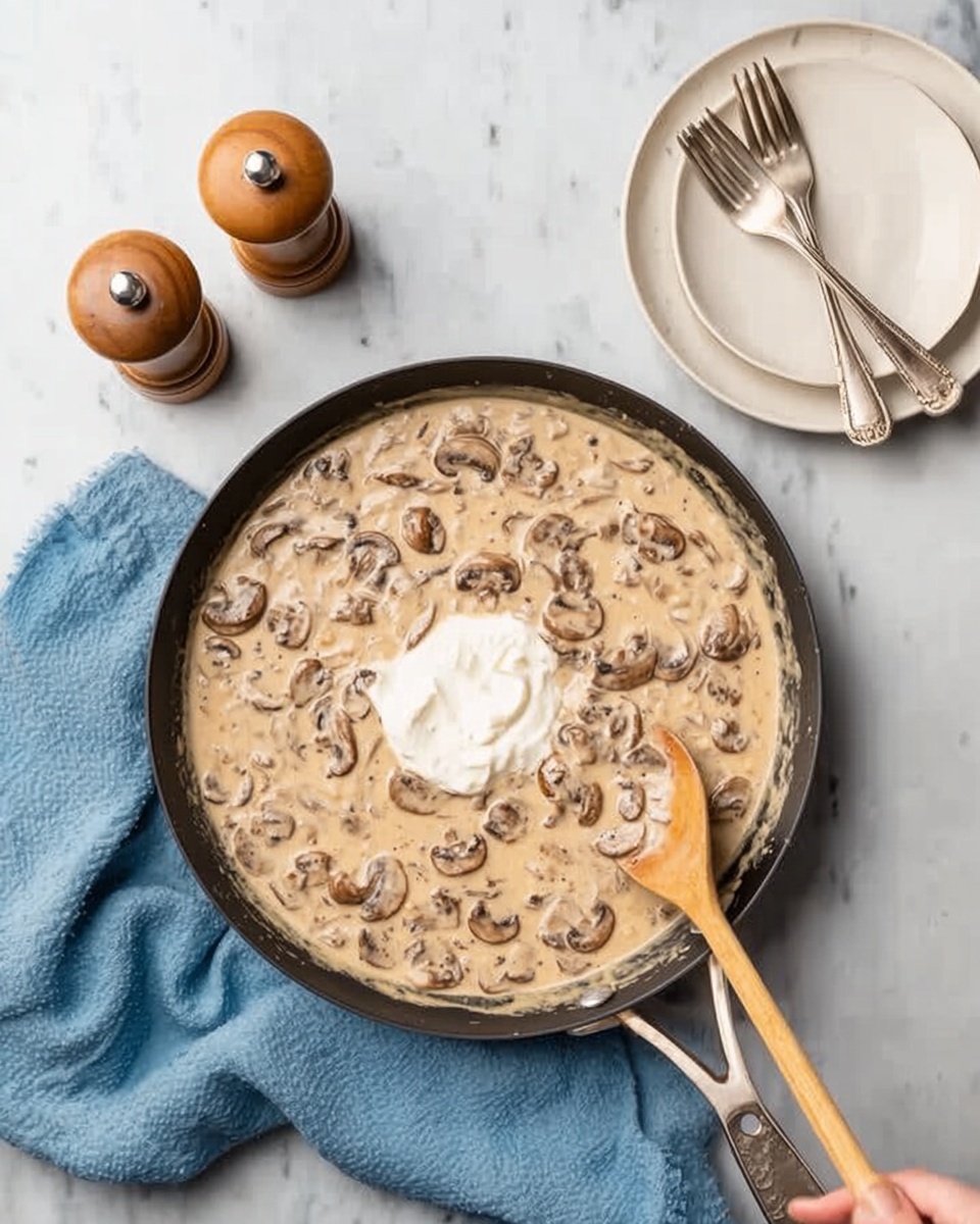 A black frying pan filled with creamy beige mushroom sauce with sliced mushrooms evenly spread throughout, topped with a dollop of white cream in the center. A woman's hand holds a wooden spoon stirring the sauce from the bottom left side of the pan. To the upper right, there is an empty white plate with two silver forks crossed on top, and to the upper left, a wooden salt and pepper shaker set on a white marbled surface. A soft blue cloth lies beneath the pan on the left side. photo taken with an iphone --ar 4:5 --v 7