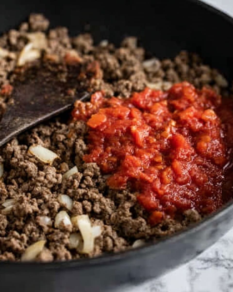 The image shows a close-up of cooked ground meat mixed with small pieces of white onion in a black pan. On one side of the pan, there is a bright red chunky tomato sauce placed on top of the meat, with a mix of tomato pieces visible. A dark wooden spoon is partially visible, resting on the meat near the sauce. The background has a white marbled texture. Photo taken with an iphone --ar 4:5 --v 7