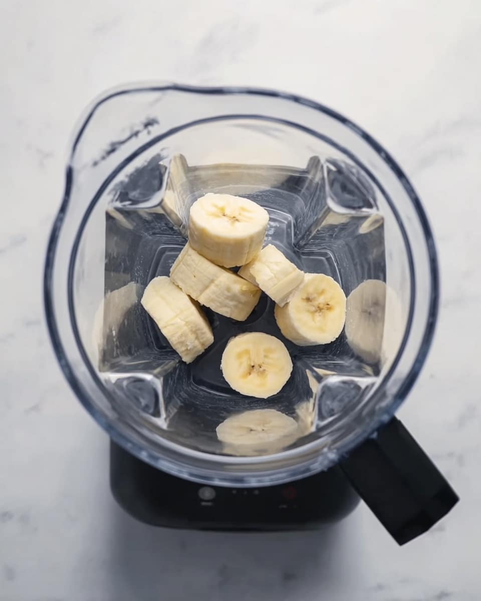 A top view of a clear blender pitcher on a white marbled surface. Inside the blender, there are four small pieces of peeled banana, placed in the center and spread slightly apart. The blender base is black, surrounding the bottom-right part of the pitcher. The overall scene is bright and clean. photo taken with an iphone --ar 4:5 --v 7