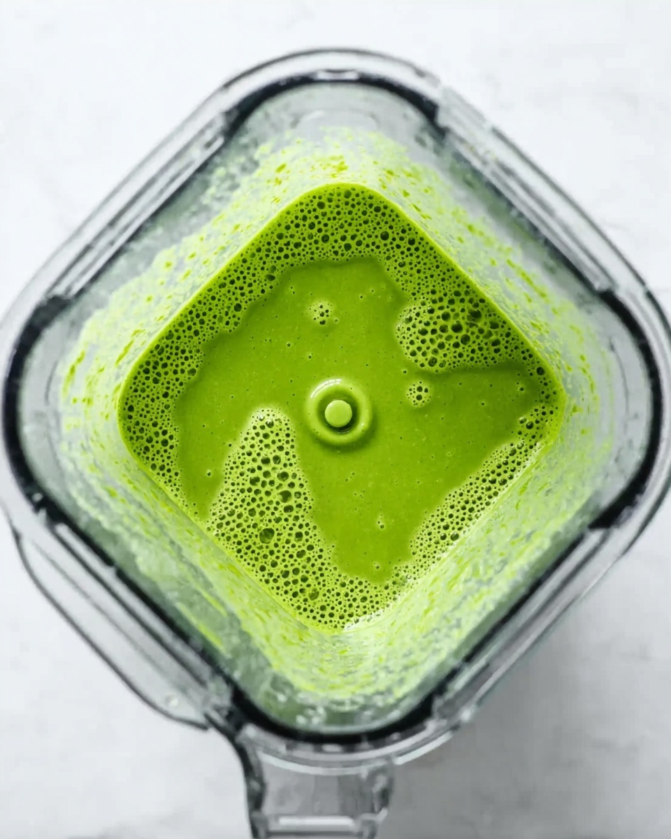 The image shows a blender from above, filled with a bright green liquid smoothie. The liquid has a thick but smooth texture, with small bubbles covering the surface. The inside of the blender is clear, showing the green mixture swirling around the blades at the bottom. Around the blender, there is a white marbled texture surface. The bright green color of the smoothie contrasts with the transparent blender walls and the clean white background. Photo taken with an iphone --ar 4:5 --v 7