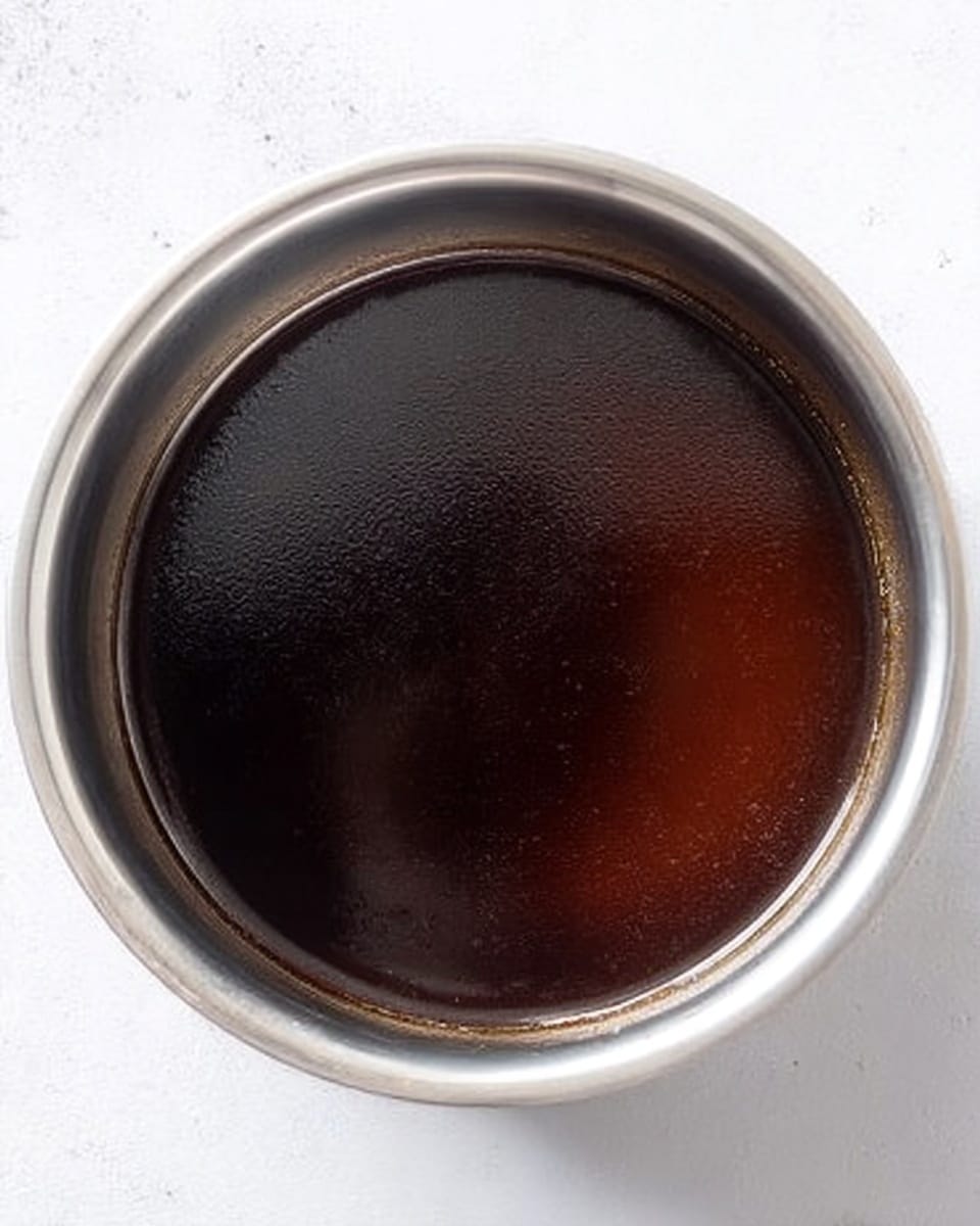 The image shows a close-up top view of a silver pot filled with dark brown, shiny liquid that looks smooth and thick. The pot sits on a white marbled surface, with the rim of the pot silver and reflecting light. There is no visible steam or other items around the pot. The focus is on the dark liquid inside, which has a glossy texture. photo taken with an iphone --ar 4:5 --v 7