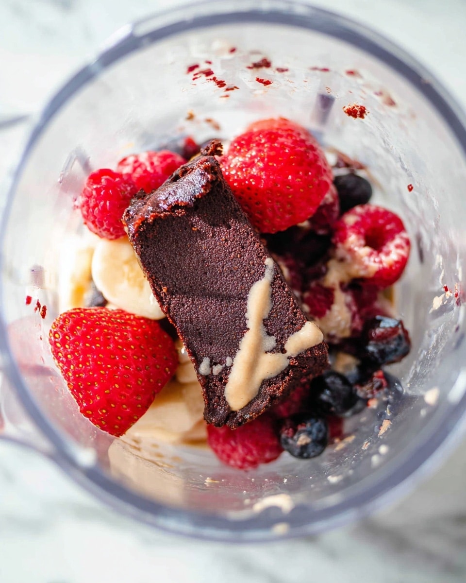 Inside a clear blender container, there is a close-up of mixed ingredients arranged in layers. At the bottom and spread thickly is a dark brown, almost black, fudgy rectangle with a rough texture. Around and partially on top of it are bright red strawberries with smooth shiny skin, smaller bright red raspberries, and a few dark blue berries. There is a light beige creamy sauce drizzled unevenly over the dark rectangle and berries. The background visible at the bottom is a white marbled surface. photo taken with an iphone --ar 4:5 --v 7