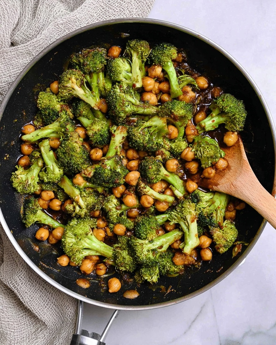 A frying pan filled with cooked broccoli and chickpeas in a dark sauce, the broccoli florets bright green with some light brown seared spots, the chickpeas golden brown and round, both mixed evenly throughout the pan. A wooden spatula rests inside the pan near the right edge. The pan sits on a white marbled surface with a textured light gray cloth in the top left corner. Photo taken with an iphone --ar 4:5 --v 7