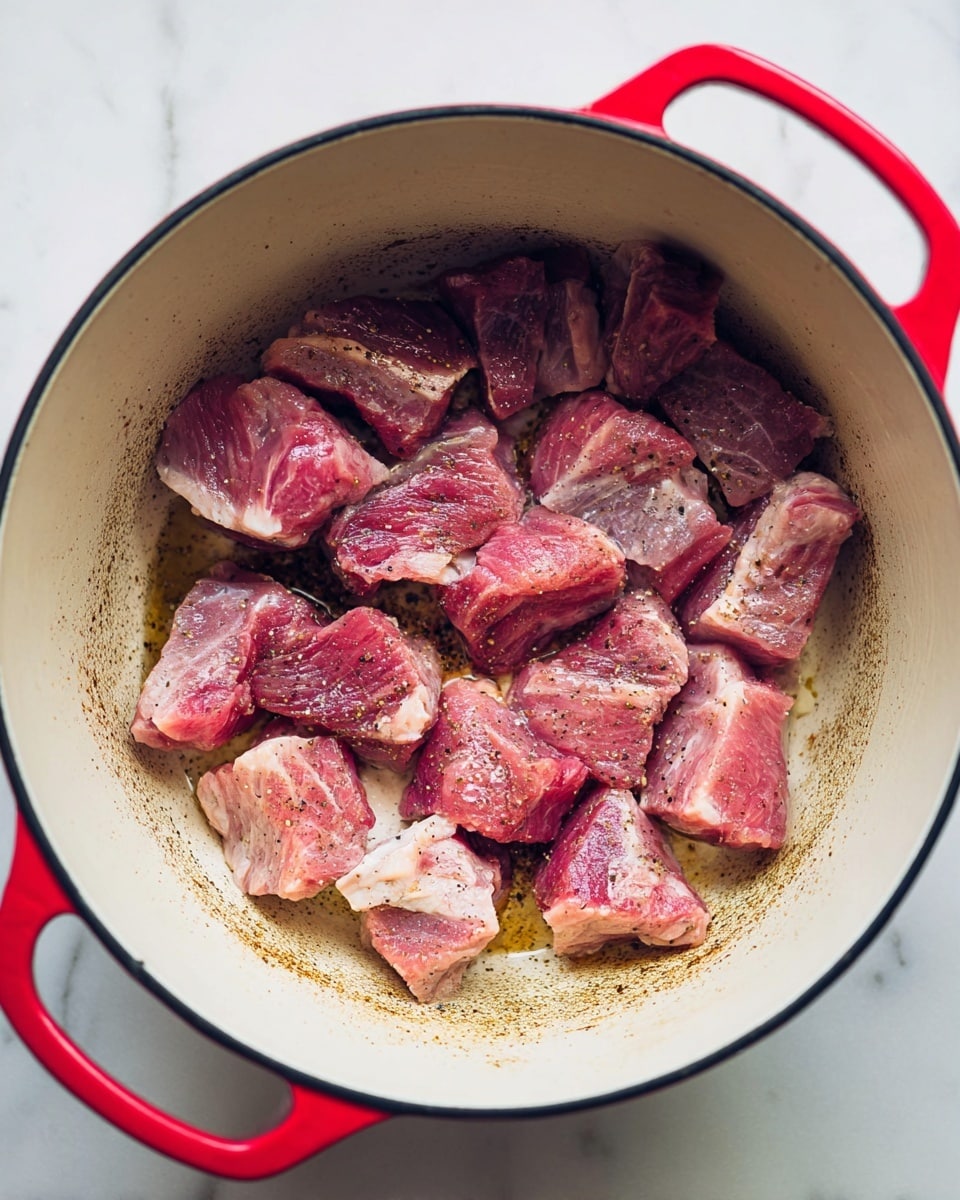 Inside a white enamel pot with a red handle, there are about twenty small pieces of raw meat being cooked. The meat is pink with some white fat and light seasoning of black pepper visible on top. The bottom of the pot shows some brown cooked marks where the meat is frying. The pot is placed on a white marbled surface. photo taken with an iphone --ar 4:5 --v 7