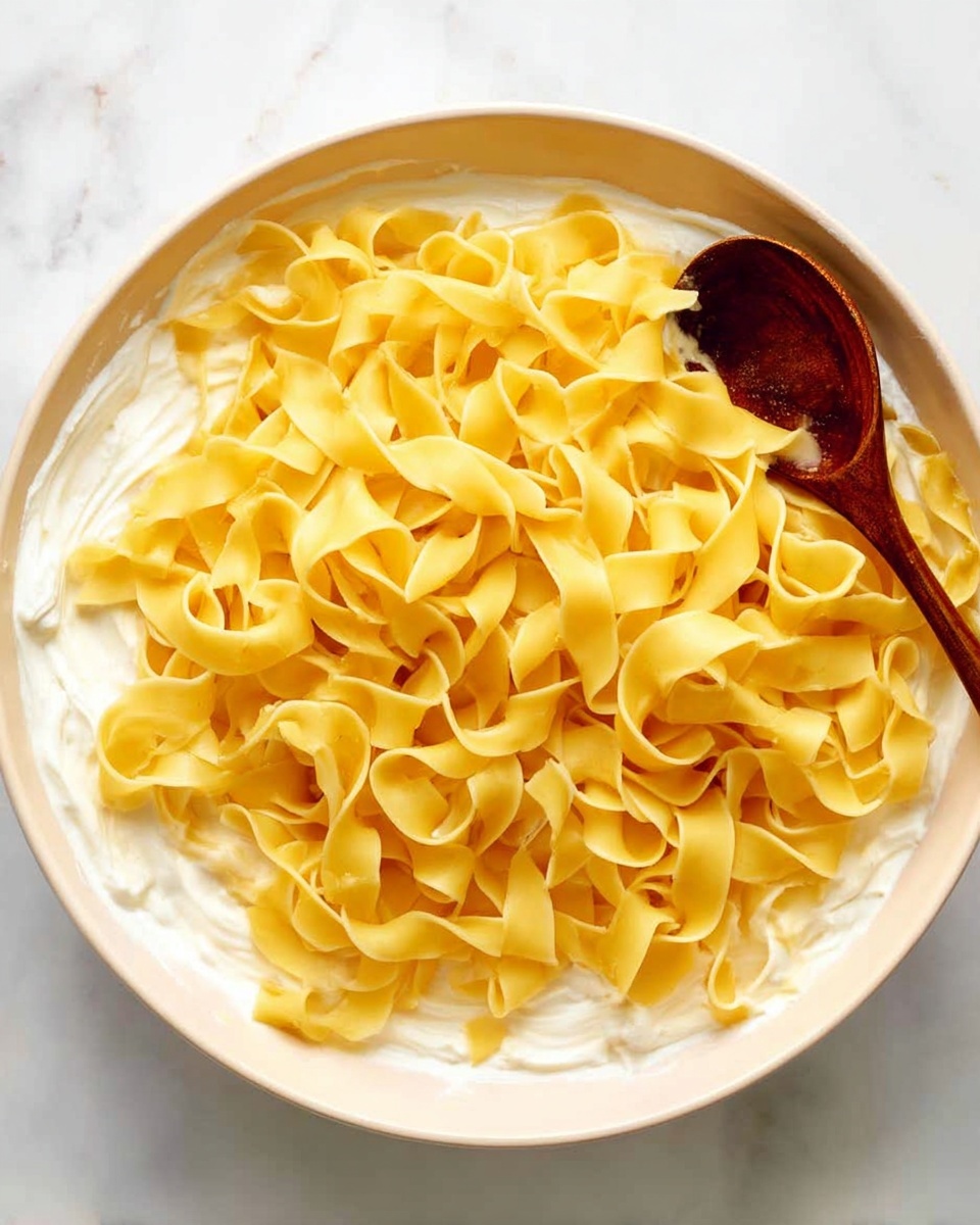 A large white bowl filled with creamy white sauce at the bottom, topped with a generous layer of light yellow wide flat noodles arranged loosely in swirls covering most of the sauce. A wooden spoon with a dark brown handle rests on the right side of the bowl, partially touching the noodles. The bowl sits on a white marbled surface. Photo taken with an iphone --ar 4:5 --v 7