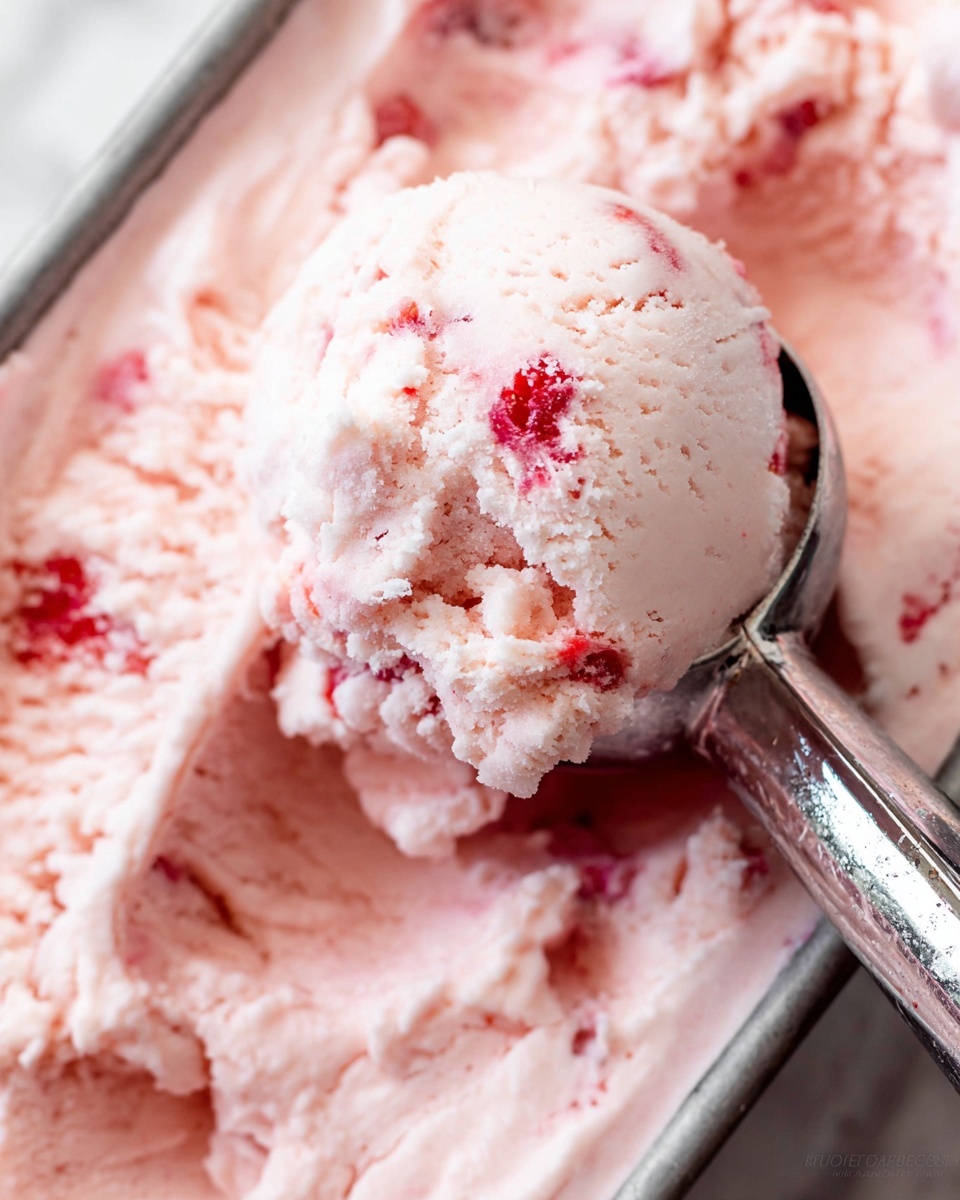 The image shows a close-up of a single scoop of light pink strawberry ice cream held by a shiny silver ice cream scoop. The ice cream has a creamy texture with visible small red strawberry pieces spread throughout. The scoop is slightly rough with soft, smooth edges, resting above a container filled with more of the same ice cream, showing uneven, swirled layers and chunks of strawberry. The background displays a white marbled surface. photo taken with an iphone --ar 4:5 --v 7