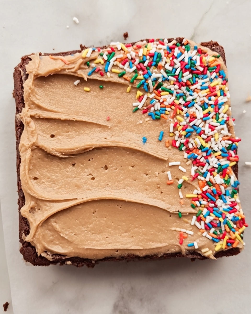 A square brownie with a thick layer of smooth, light brown frosting spread evenly on top, showing soft, wavy texture marks made by a spatula. The frosting is decorated with colorful sprinkles mostly concentrated on the upper right corner, with colors including red, white, yellow, blue, green, and orange. The brownie sits on a white marbled surface. photo taken with an iphone --ar 4:5 --v 7