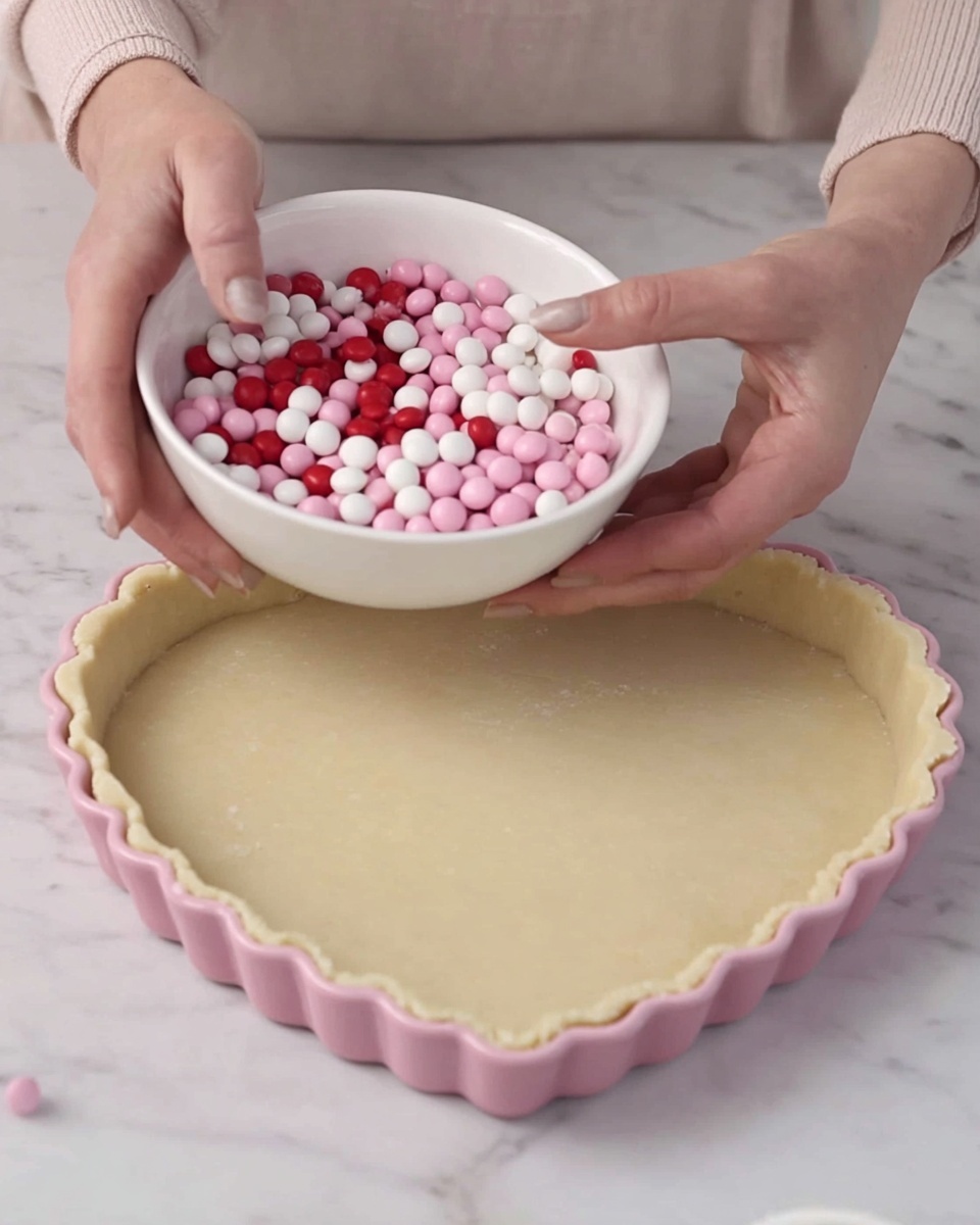 A woman's hand is holding a white bowl filled with small round candies in shades of pink, white, and red. The other woman's hand is placing a single white candy on the edge of a dough-covered tart that is in a pink heart-shaped tart pan with a fluted edge. The dough is light beige and smooth, covering the entire tart pan. The scene is set on a white marbled surface, and the background shows a light kitchen setting. photo taken with an iphone --ar 4:5 --v 7