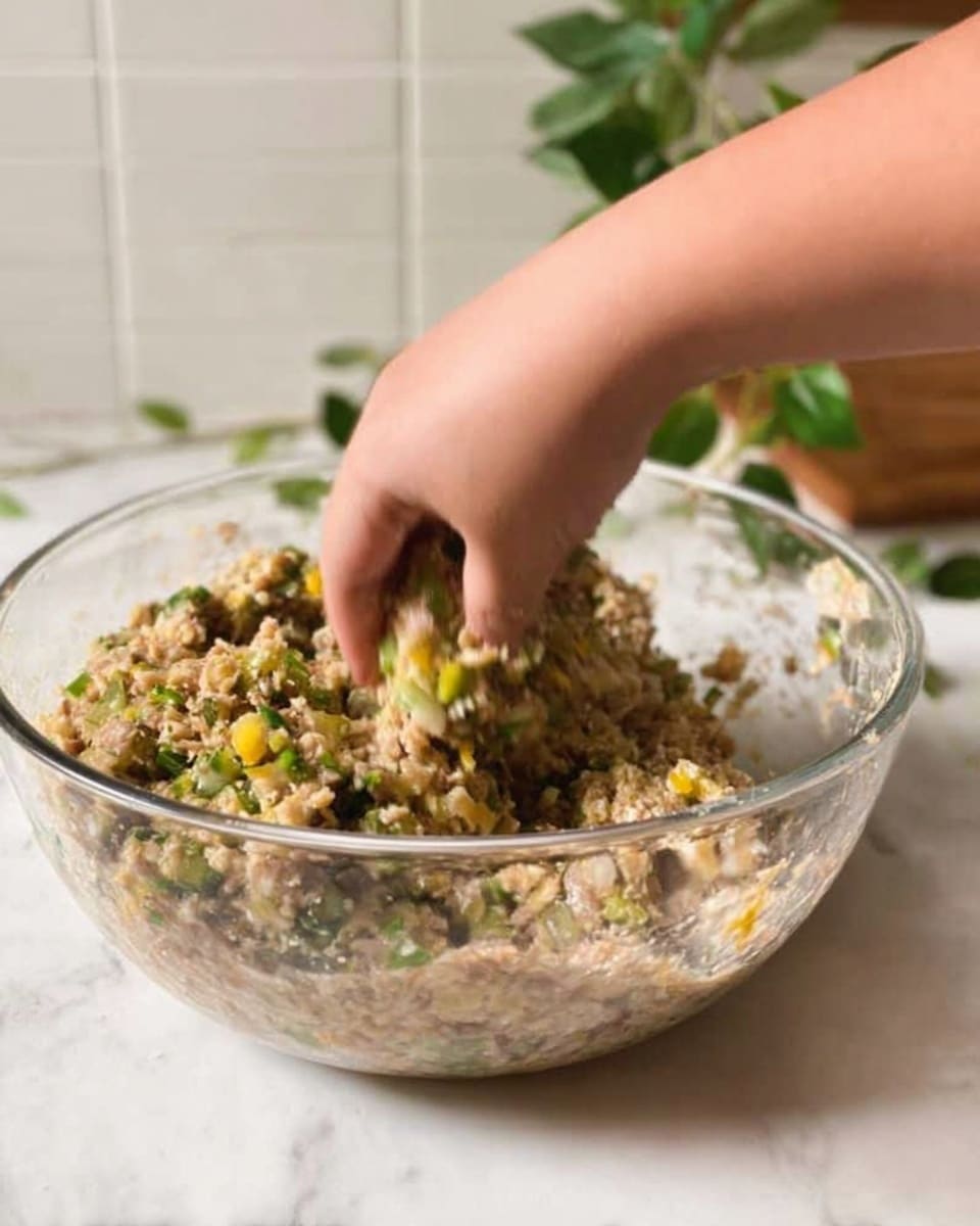 A clear glass bowl filled with a mixture of small chopped vegetables and grains in light brown and green colors, being mixed by a woman's hand. The mixture looks moist with visible bits of green herbs and yellow pieces, showing a sticky texture. The bowl is placed on a white marbled surface, with green leaves and a white tiled wall faintly visible in the background. Photo taken with an iphone --ar 4:5 --v 7