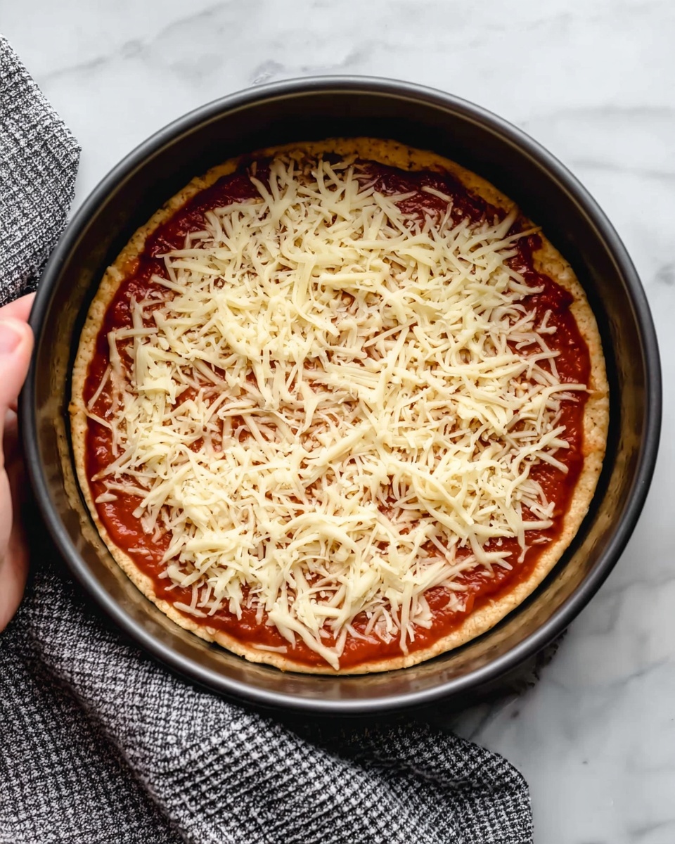 A round black cake pan holds a three-layer pizza. The bottom layer is golden brown pizza dough, smooth and even. On top is a thick layer of red tomato sauce spread close to the edges, with a shiny, slightly wet texture. The top layer is a generous pile of finely shredded pale yellow cheese covering the sauce fully, with some strands loosely spread. A woman's hand holds the cake pan handle on the right side. The pan is placed on a white marbled surface with a grey and white checkered cloth nearby. photo taken with an iphone --ar 4:5 --v 7