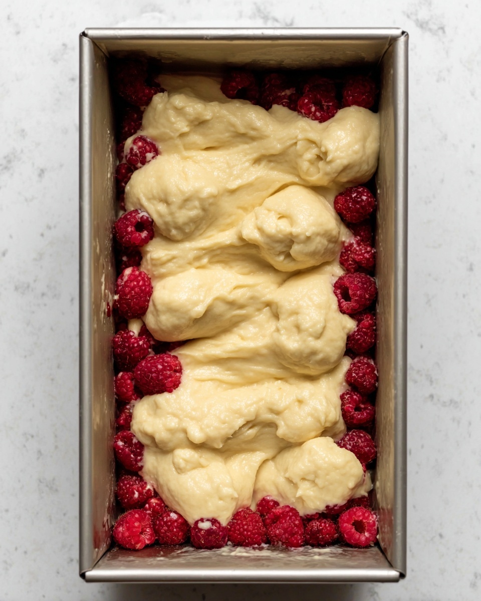 The image shows a rectangular metal baking pan filled with a layer of whole raspberries spread evenly on the bottom. On top of the raspberries, there is a thick, pale yellow batter dolloped in uneven mounds, not yet spread out. The pan is placed on a white marbled surface. Photo taken with an iphone --ar 4:5 --v 7