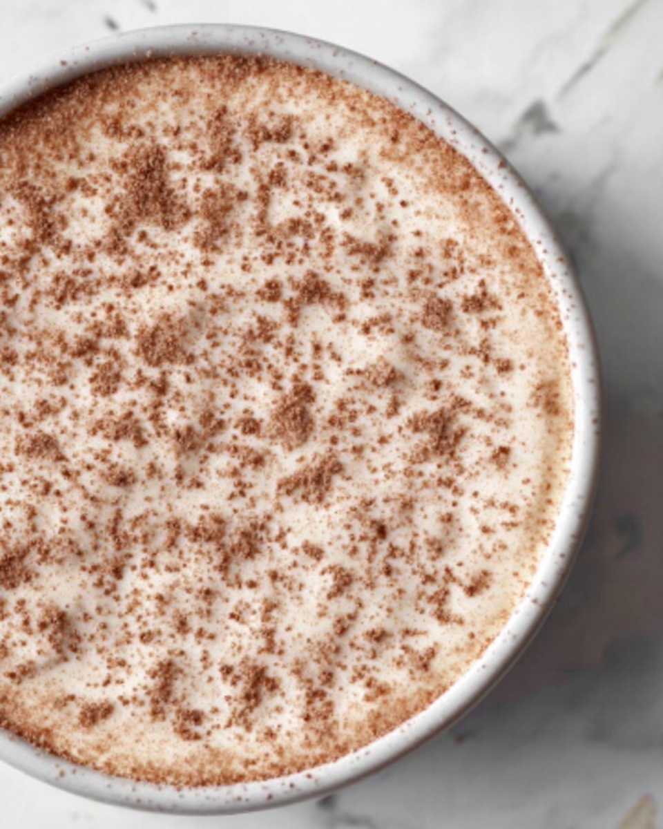 A close-up image of a round dessert in a white bowl on a white marbled surface. The dessert has a smooth, creamy top layer with a light brown color sprinkled evenly with a fine layer of brown powder. The texture looks soft and slightly fluffy with evenly distributed tiny specks across the surface. The bowl's edge is visible, showing it is neatly filled to just below the rim. The lighting highlights the subtle shadows and details of the creamy texture. photo taken with an iphone --ar 4:5 --v 7
