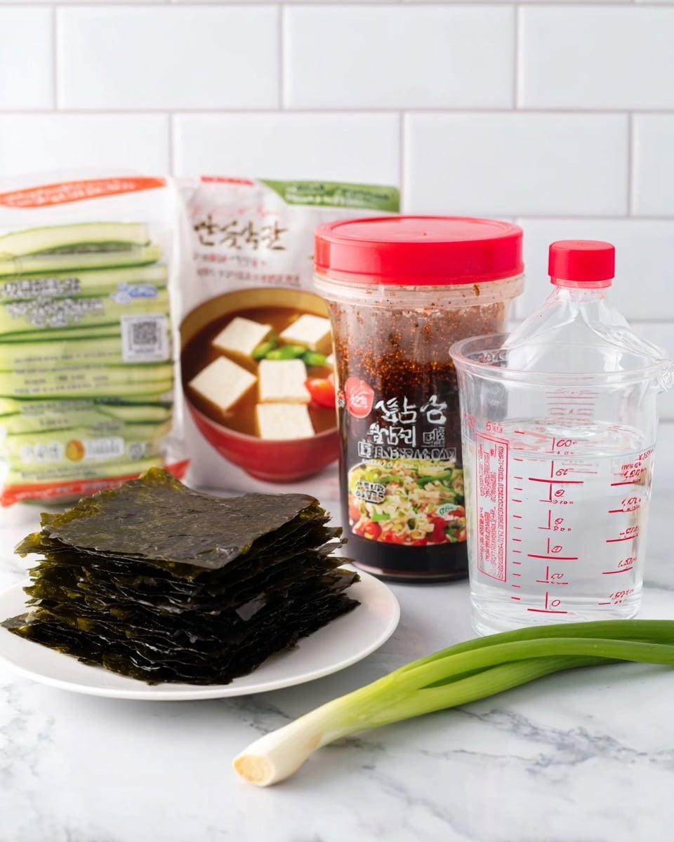 The image shows a white plate with dark green, dried seaweed sheets stacked on it in the front left. Behind it to the left is a white marbled surface with a package featuring tofu, cucumbers, and tomatoes visible in the background. In the middle is a clear plastic container of red soybean paste with a red cap and a label showing soup with tofu and greens. On top of the paste container sits a small clear bottle of red seasoning with a red cap. To the right of the paste is a clear measuring cup filled halfway with water, marked with red measurement lines. A fresh green onion lies horizontally in front of the soybean paste and measuring cup. The whole scene is set against a clean white tiled wall and a white marbled surface photo taken with an iphone --ar 4:5 --v 7