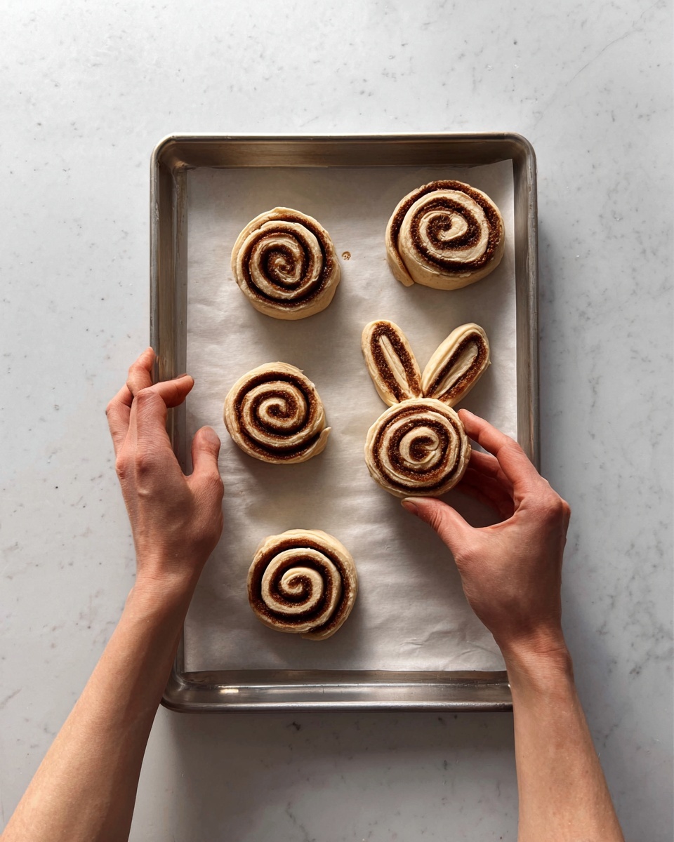 A metal tray lined with white parchment paper holds five pieces of rolled dough with a cinnamon swirl. Four of the rolls are round with visible spiral layers in light tan dough and darker brown cinnamon filling. One roll at the top is shaped like a bunny face with two ears, featuring the same spiral swirl pattern. A woman's two hands are positioned over the tray, with the left hand holding a round cinnamon roll and the right hand poised above it. The whole setup is on a white marbled surface. Photo taken with an iphone --ar 4:5 --v 7