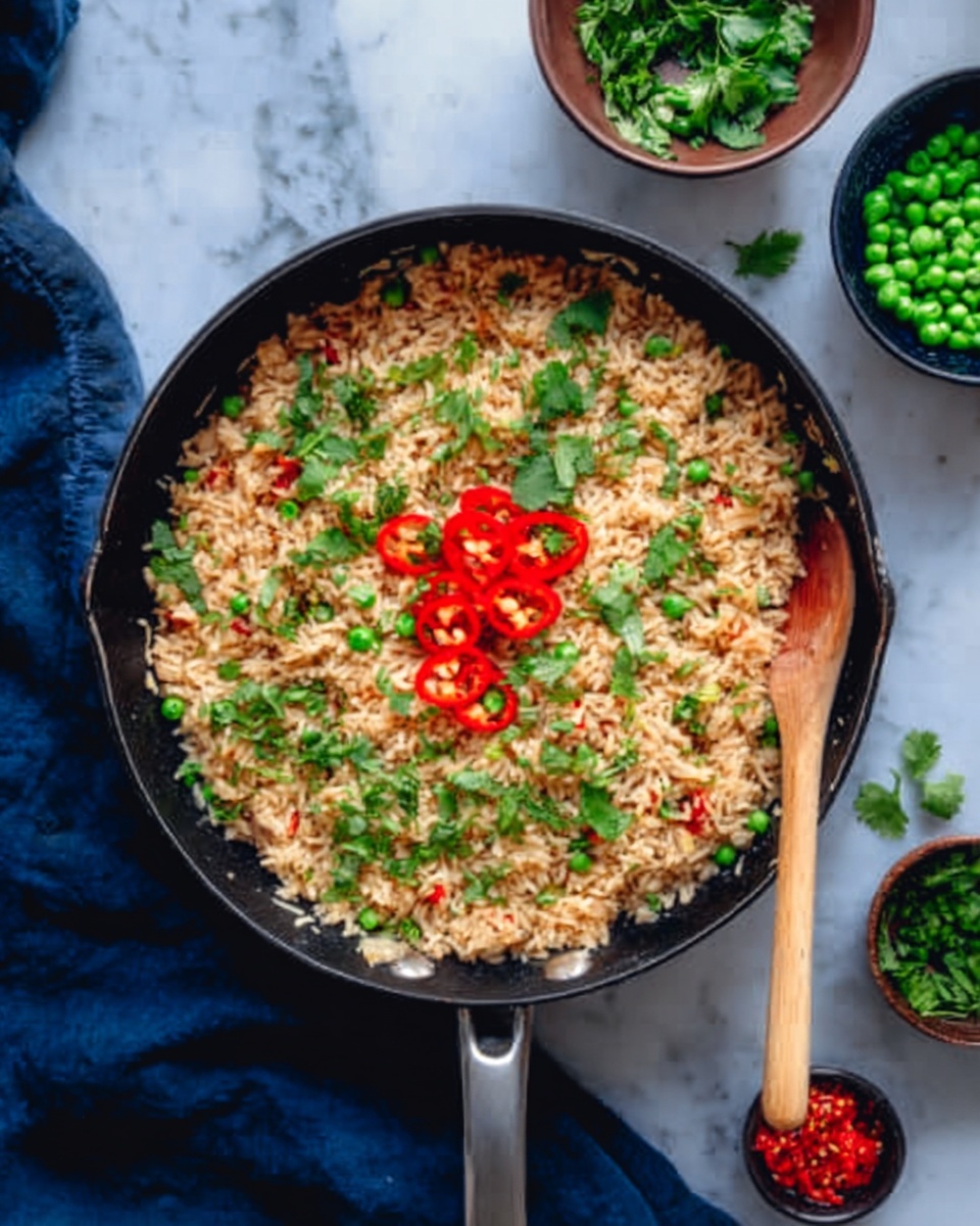 A black pan placed on a white marbled surface holds a cooked rice dish with light brown grains. On top of the rice, there are slices of bright red chili peppers scattered in the middle, some green herbs like cilantro spread around, and small bits of green peas mixed throughout. A wooden spoon rests inside the pan on the right side. Nearby, small bowls contain chopped green herbs, green peas, and red chopped ingredients, all on the white marbled surface. A navy blue cloth is partially visible under the pan. Photo taken with an iphone --ar 4:5 --v 7