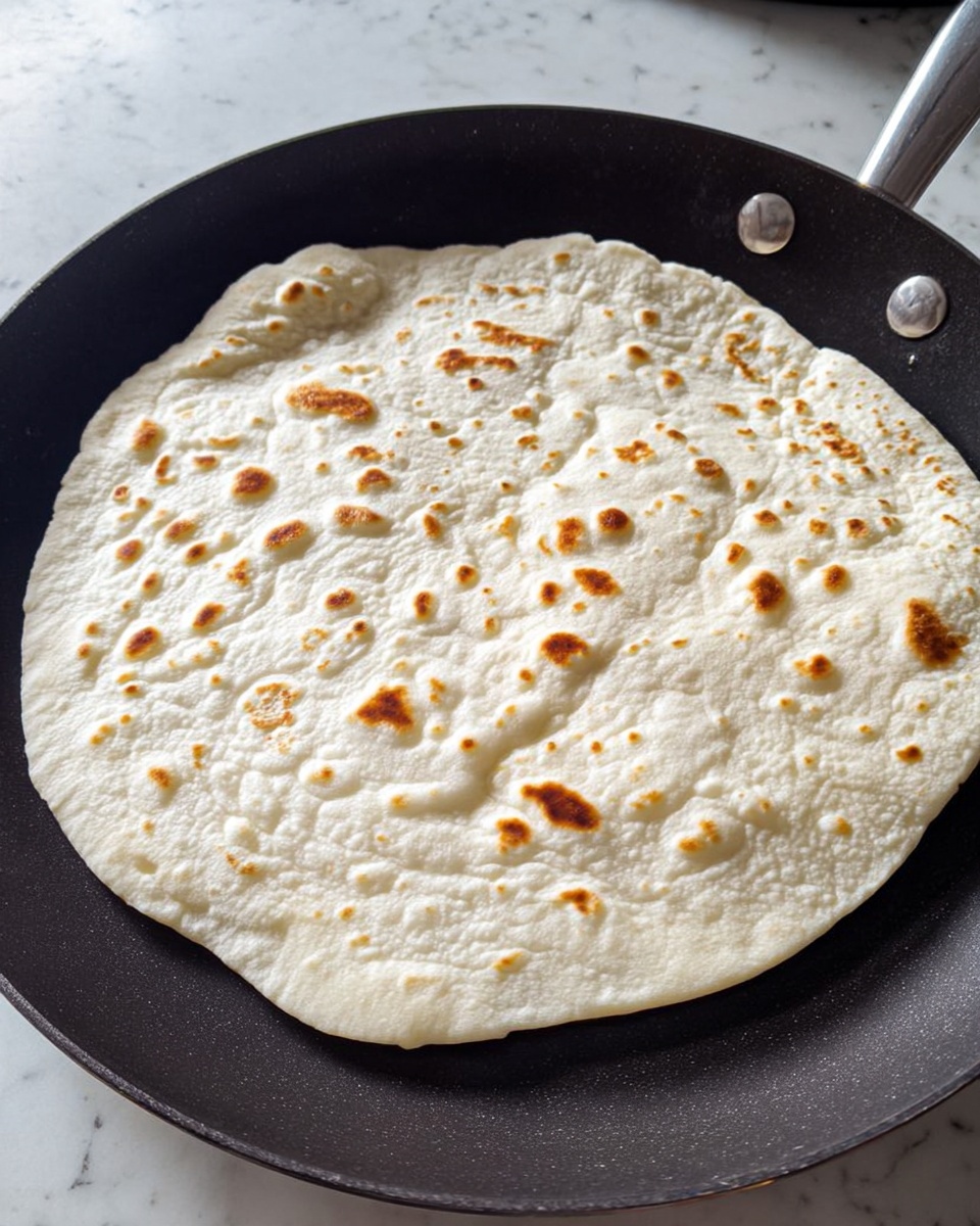 A thin flatbread with a light white texture and small brown toasted spots is cooking on a black pan. The flatbread has a few raised bubbles, adding a puffed texture to some parts of it. The pan sits on a white marbled surface with a small round silver handle visible. The flatbread covers most of the pan's surface and looks soft yet slightly crispy around the edges. Photo taken with an iphone --ar 4:5 --v 7