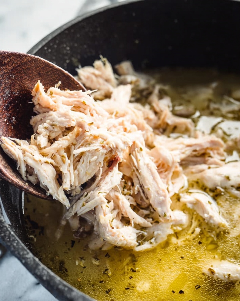 A close-up shot shows shredded light tan chicken being moved from a white textured plate into a black pot with yellowish broth inside. The shredded chicken has a soft and fibrous texture, loosely piled on the plate and falling into the broth below. A dark wooden spoon is partially visible, angled under some of the shredded chicken. The black pot has a worn, matte surface and the broth inside is speckled with light herbs and small oil droplets, giving a slightly oily, reflective surface. The background features a white marbled texture. photo taken with an iphone --ar 4:5 --v 7