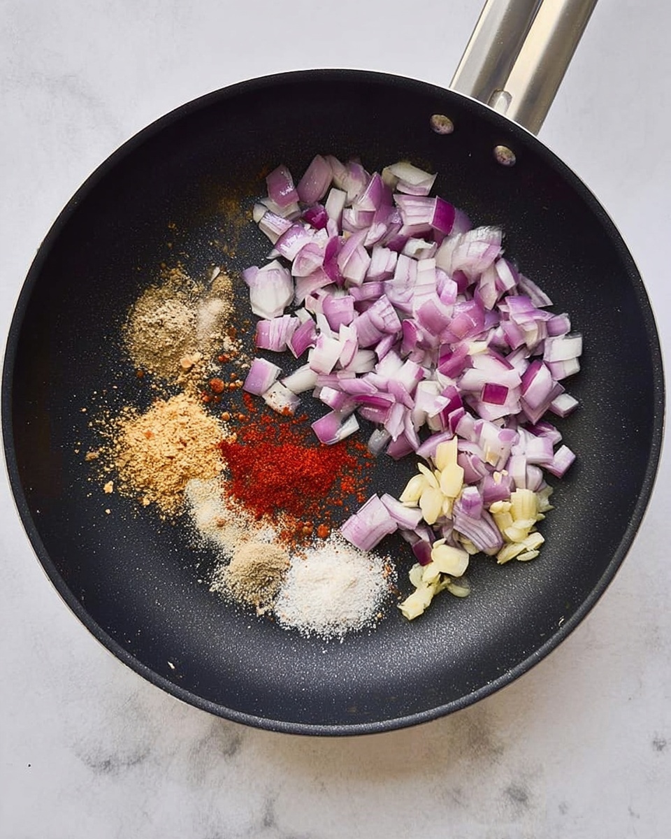 A black frying pan with a silver handle is shown from above, resting on a white marbled surface. Inside the pan, there is a mix of small chopped pieces of purple and white onions scattered mainly on the right side. In the center right, there are small chunks of light yellow garlic. On the left side of the pan, four different powders are arranged in small piles: a light brown powder on top, a beige powder below it, a brighter red powder in the middle, and a small amount of white powder at the bottom. The textures range from crunchy onion pieces to soft powder. Photo taken with an iphone --ar 4:5 --v 7