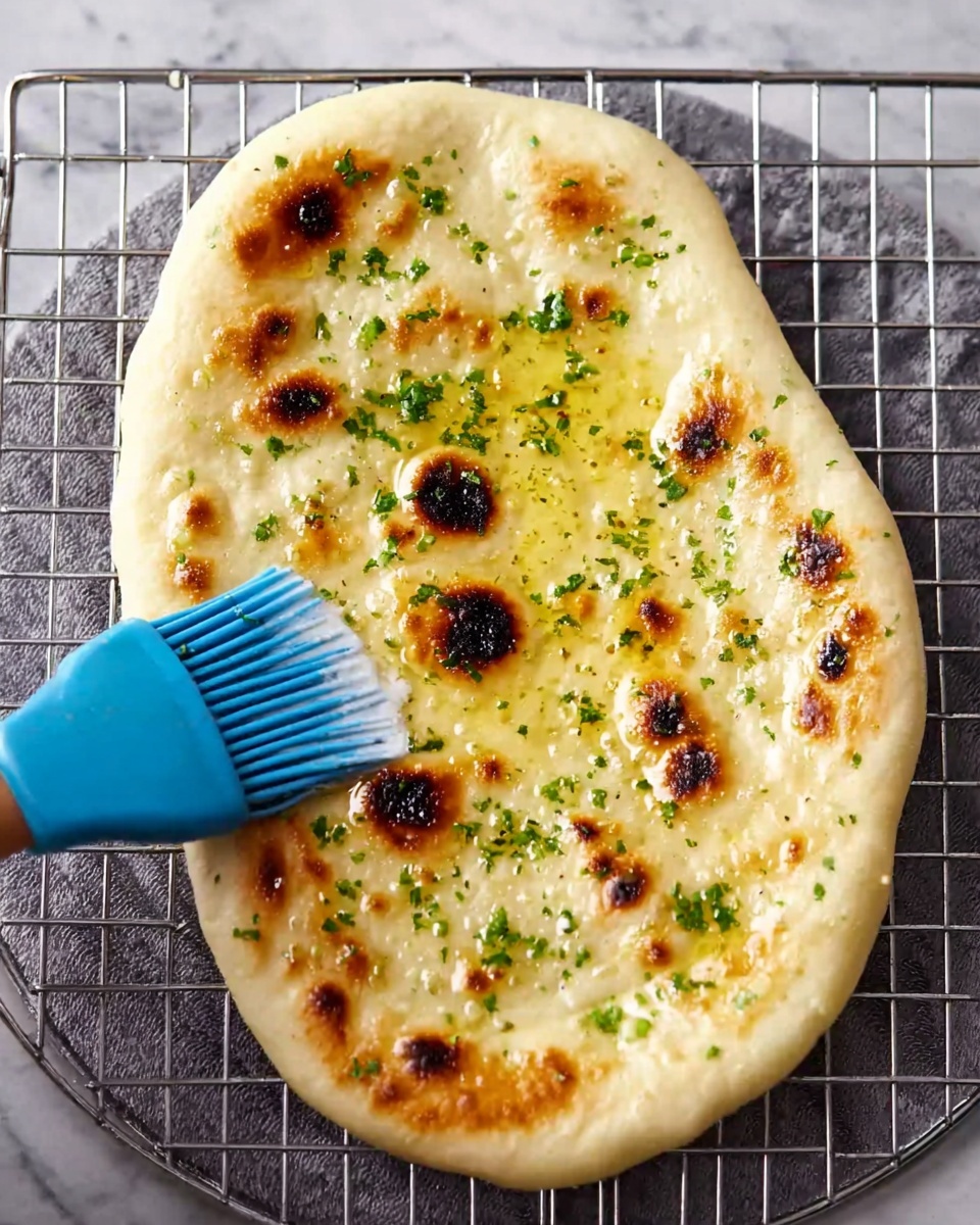 The image shows a piece of naan bread on a round wire rack, placed over a white marbled surface. The naan is oval-shaped with golden brown spots scattered across its surface, indicating it is freshly cooked. The texture looks soft with some bubbling and light charring. On top of the naan, there are small green herb pieces sprinkled evenly. A woman's hand is holding a blue brush, spreading a shiny layer of melted butter across the naan, giving it a glossy finish. photo taken with an iphone --ar 4:5 --v 7