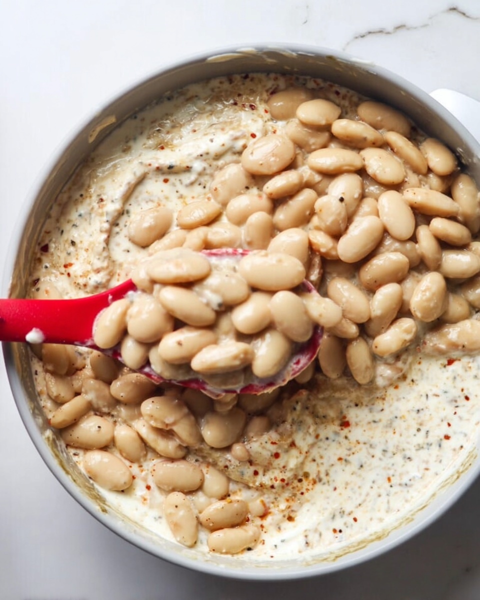 The image shows a close-up of a bowl with two main layers. The bottom layer is white creamy sauce with specks of seasoning, giving it a slightly speckled texture. On top, there is a thick layer of pale beige beans covering the creamy base. A red spoon is partially dipped into the dish, lifting some beans. The bowl is white and it is placed on a white marbled surface. Photo taken with an iphone --ar 4:5 --v 7