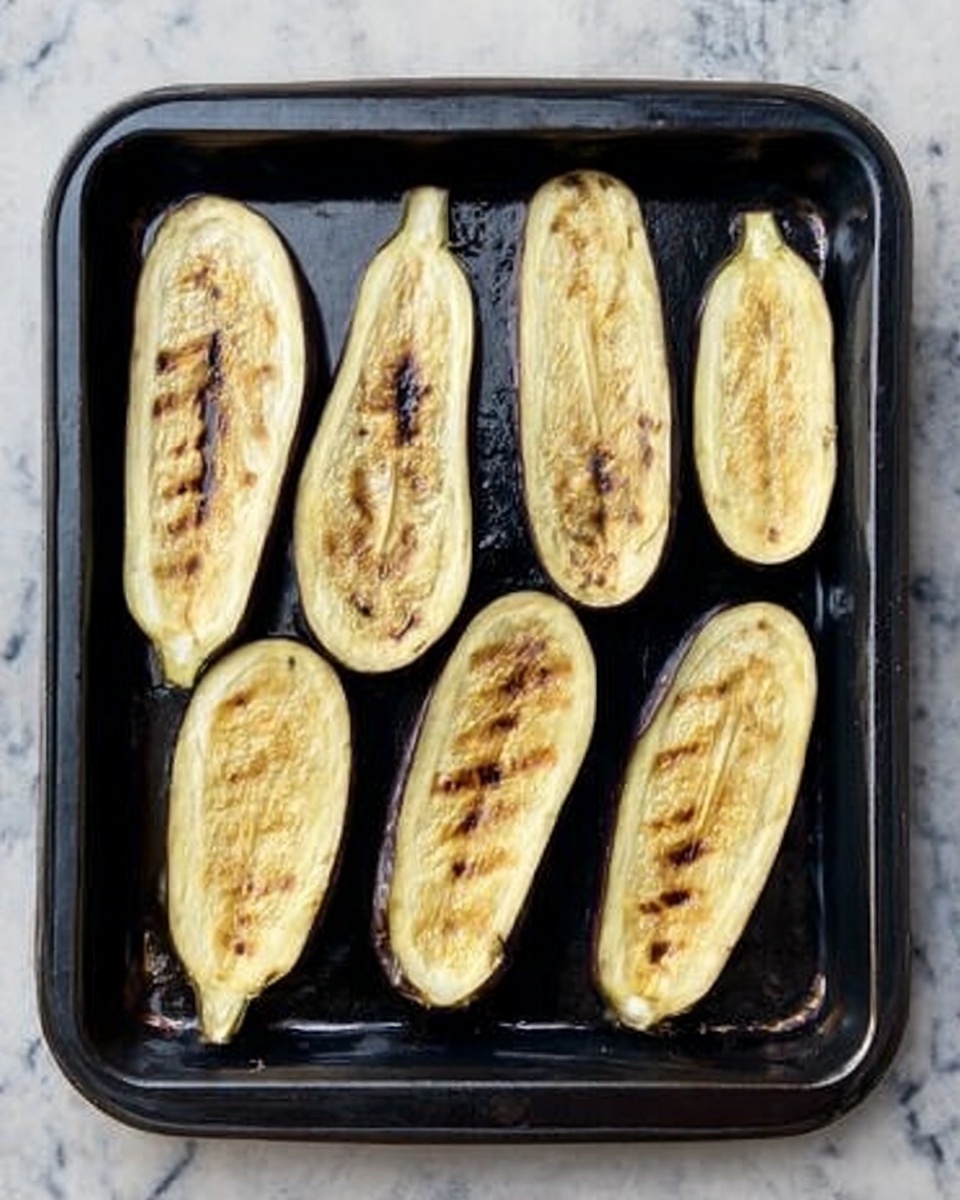 The image shows a black baking tray with nine slices of roasted eggplant evenly spread out. Each slice is a pale yellowish color with brown grill marks and slightly crispy edges, showing a soft texture inside. The tray sits on a white marbled surface, adding contrast to the dark tray and light eggplants. The eggplant slices vary slightly in size, arranged neatly in two rows with some space between them. photo taken with an iphone --ar 4:5 --v 7