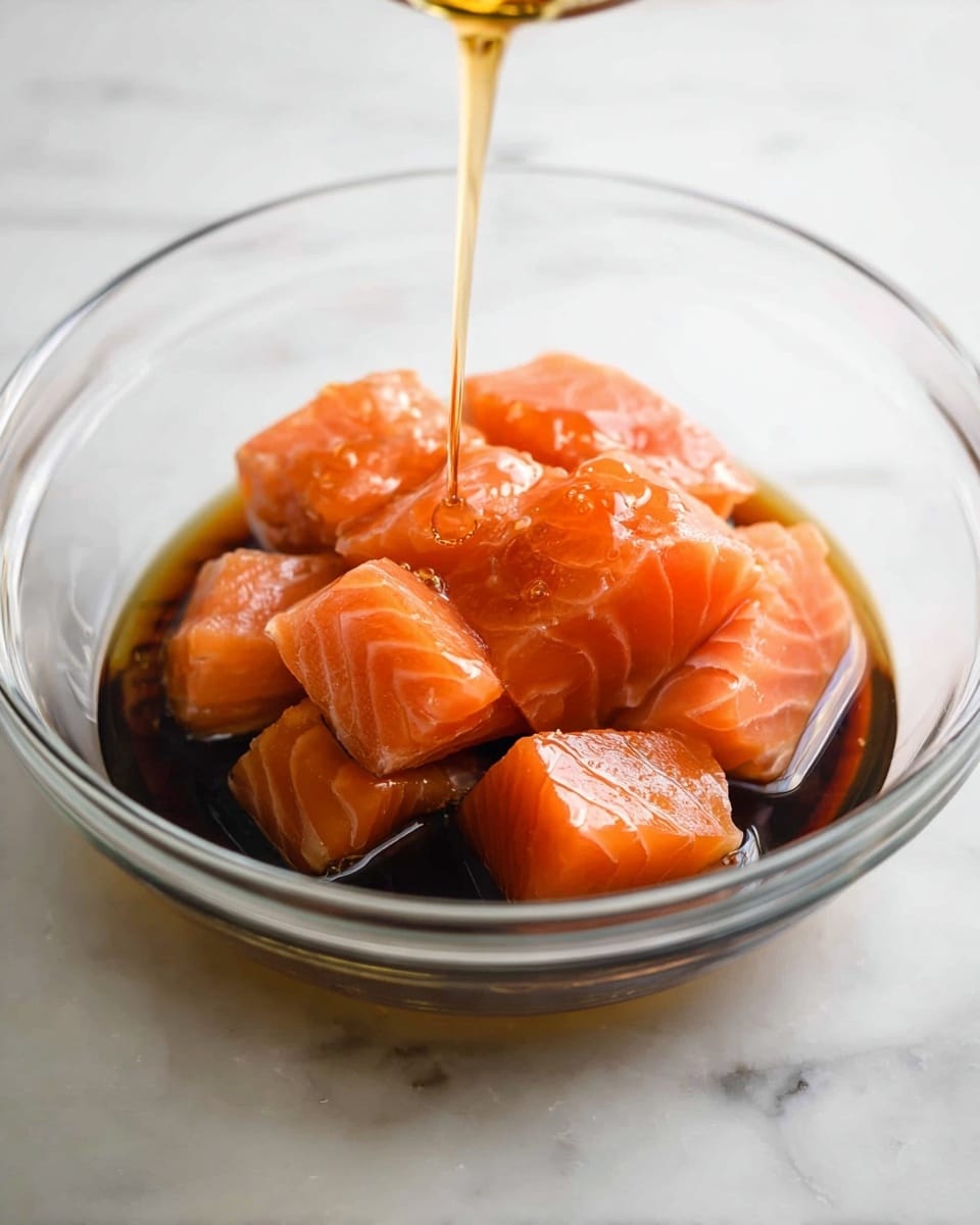 A clear round glass bowl holds several pieces of raw salmon cut into thick rectangular chunks with a bright orange-pink color and soft texture. The salmon pieces rest in a dark brown soy sauce that almost fills the bottom of the bowl, creating a glossy, wet appearance. Above the bowl, a thin golden liquid is being slowly poured, forming a smooth stream that catches the light, adding shine to the salmon. The bowl is placed on a white marbled surface that adds a clean, natural background to the scene. photo taken with an iphone --ar 4:5 --v 7