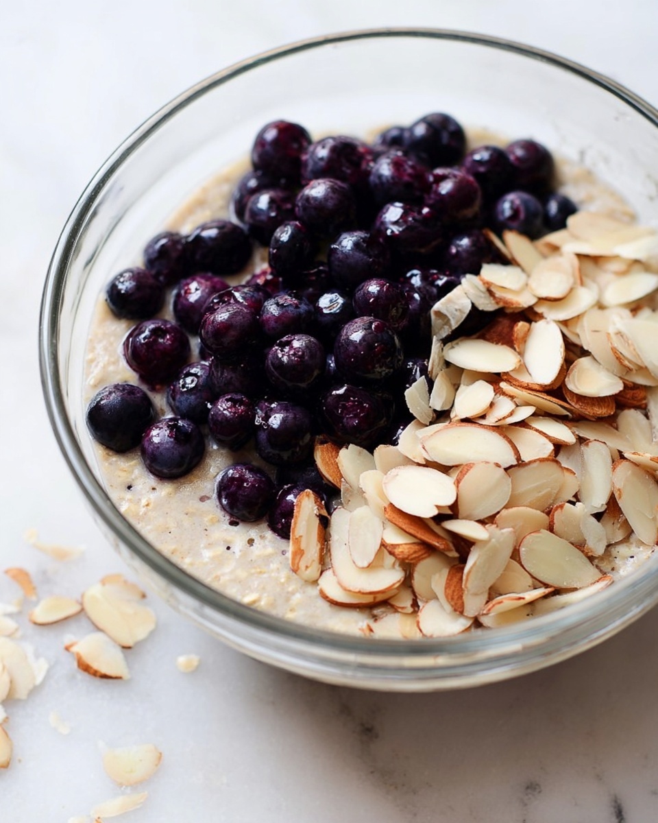 A clear glass bowl filled with a creamy beige oatmeal mixture as the base layer. On top, there is a layer of dark purple whole blueberries, clustered mainly on one side, with their smooth texture visible. Next to the blueberries, a layer of light brown sliced almonds is scattered, some overlapping the blueberries slightly. The bowl is placed on a white marbled surface, with a few stray almond slices nearby. photo taken with an iphone --ar 4:5 --v 7