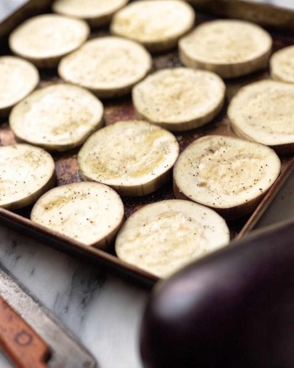 The image shows a close-up of a metal baking tray filled with light cream-colored round slices of eggplant, spread evenly and sprinkled with black pepper or seasoning. The slices have a soft, smooth texture with some light browning on the edges. In the foreground, a dark purple eggplant and a wooden-handled knife lie on a white marbled surface, slightly out of focus. The scene is simple and clean, highlighting the raw slices prepared for cooking. photo taken with an iphone --ar 4:5 --v 7