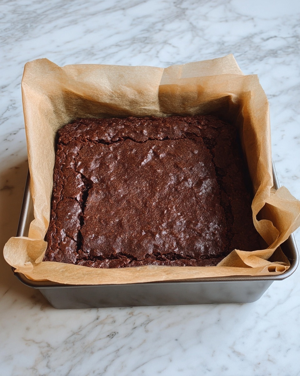The image shows a baked brownie in a square metal pan lined with light brown parchment paper that folds over the edges. The brownie has a dark brown surface with a slightly cracked texture in the middle and parts of the edges. The pan sits on a white marbled surface. photo taken with an iphone --ar 4:5 --v 7