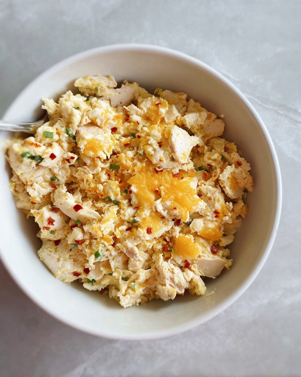A white bowl filled with a mixture of light beige fluffy scrambled eggs and small pieces of melted orange cheese spread evenly on top, with tiny green and red bits scattered throughout. The bowl rests on a white marbled surface. photo taken with an iphone --ar 4:5 --v 7