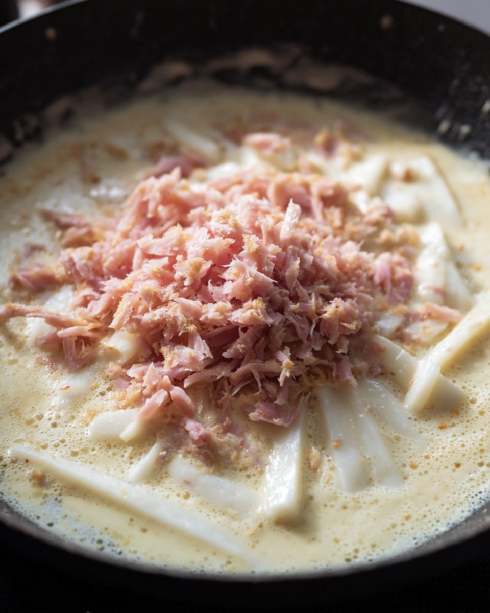 A close-up view of a dark pan filled with bubbling creamy white sauce as the base layer, with white, thick, stick-shaped pieces spread evenly inside the sauce. On top, there is a heap of finely shredded pinkish meat placed in the center, adding texture contrast. The sauce looks smooth and slightly thick, and the meat bits have a tender, moist look to them, all cooking together. Photo taken with an iphone --ar 4:5 --v 7