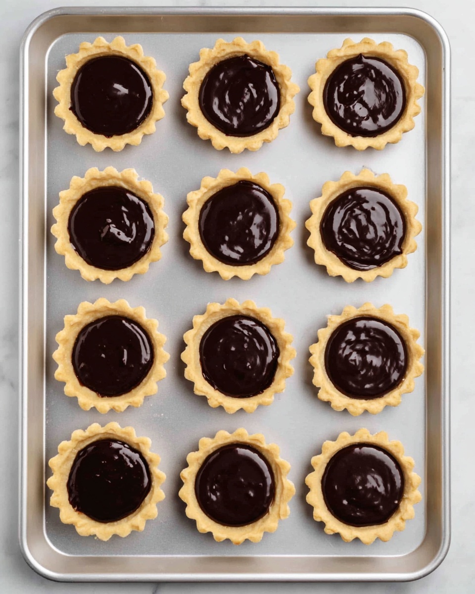 The image shows twelve small tartlets arranged in a 3x4 grid on a silver baking tray. Each tartlet has a light golden, scalloped-edged crust filled with smooth, dark chocolate ganache, which has a shiny, slightly uneven surface. The tray rests on a white marbled background, enhancing the contrast between the rich chocolate filling and the pale crust. The tartlets appear evenly spaced and the whole setup looks clean and organized. photo taken with an iphone --ar 4:5 --v 7